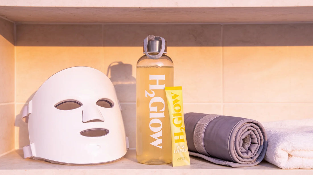 H2Glow water bottle and Fresh Lemonade stick pack on a bathroom shelf beside an LED face mask, a rolled sauna blanket and a towel