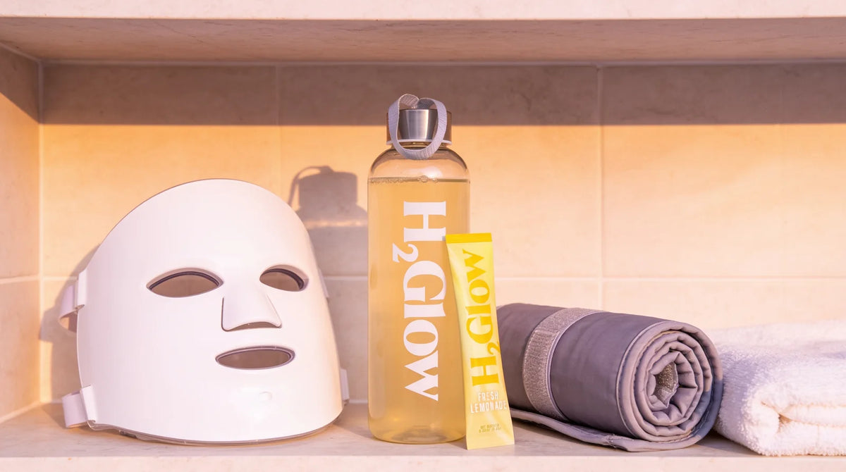 H2Glow water bottle and Fresh Lemonade stick pack on a bathroom shelf beside an LED face mask, a rolled sauna blanket and a towel