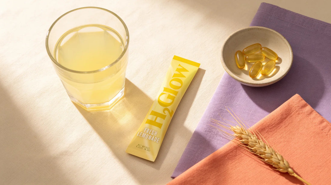 H2Glow Fresh Lemonade stick pack on cream linen beside a glass of lemon drink, a dish of softgel capsules and a sprig of wheat