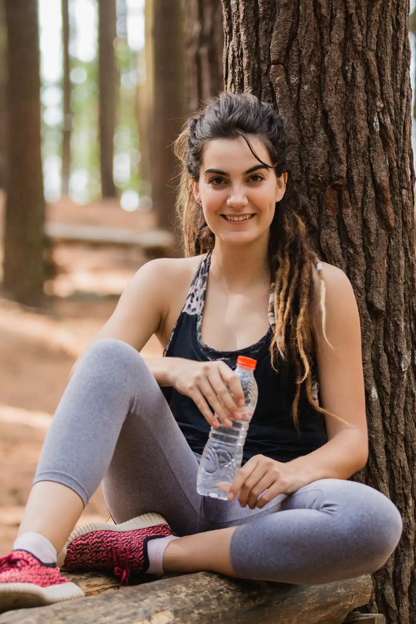 Woman with long dreadlocks sits by a tree trunk holding a clear water bottle in a sunlit park.