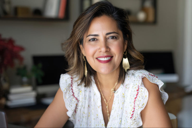 Woman in a light floral blouse smiles at the camera, wearing gold earrings in a softly blurred living room.