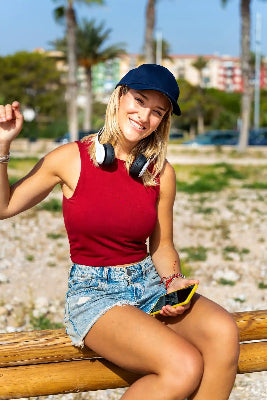 Smiling woman in a red tank top and blue cap sits on a wooden bench, wearing headphones outdoors.