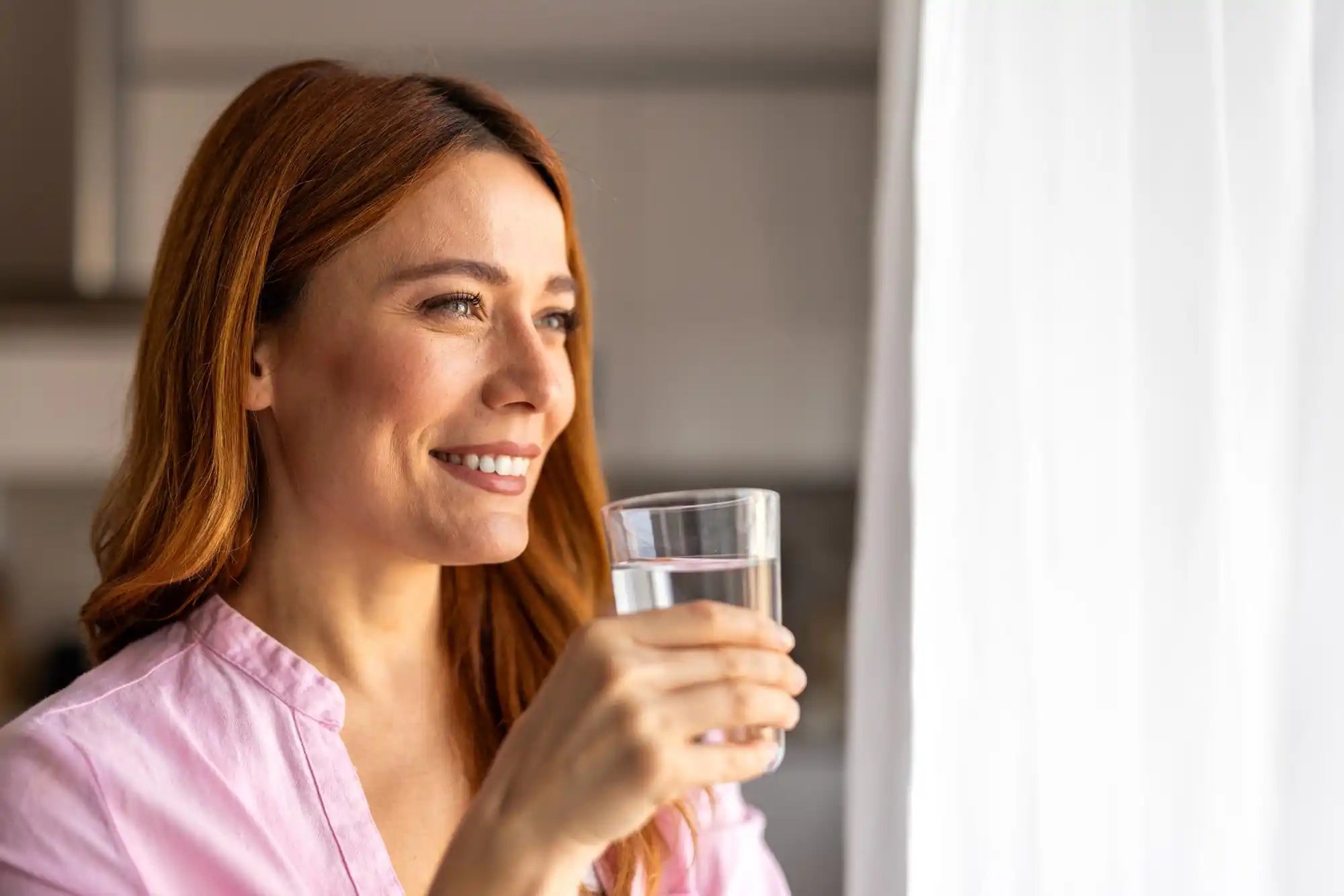 Woman holding a clear glass and smiling softly, her auburn hair and pink shirt in warm window light
