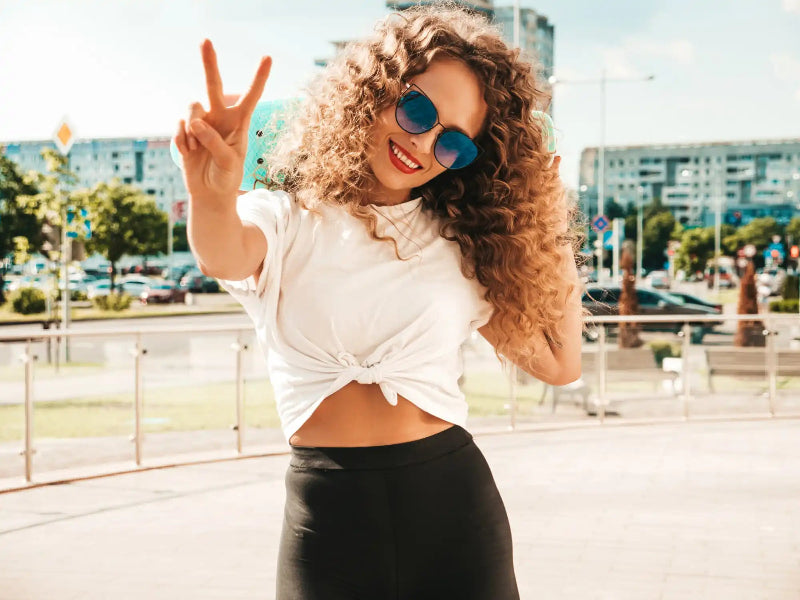 Woman with curly hair and blue mirrored sunglasses waves at a sunny rooftop, with a city skyline behind her.