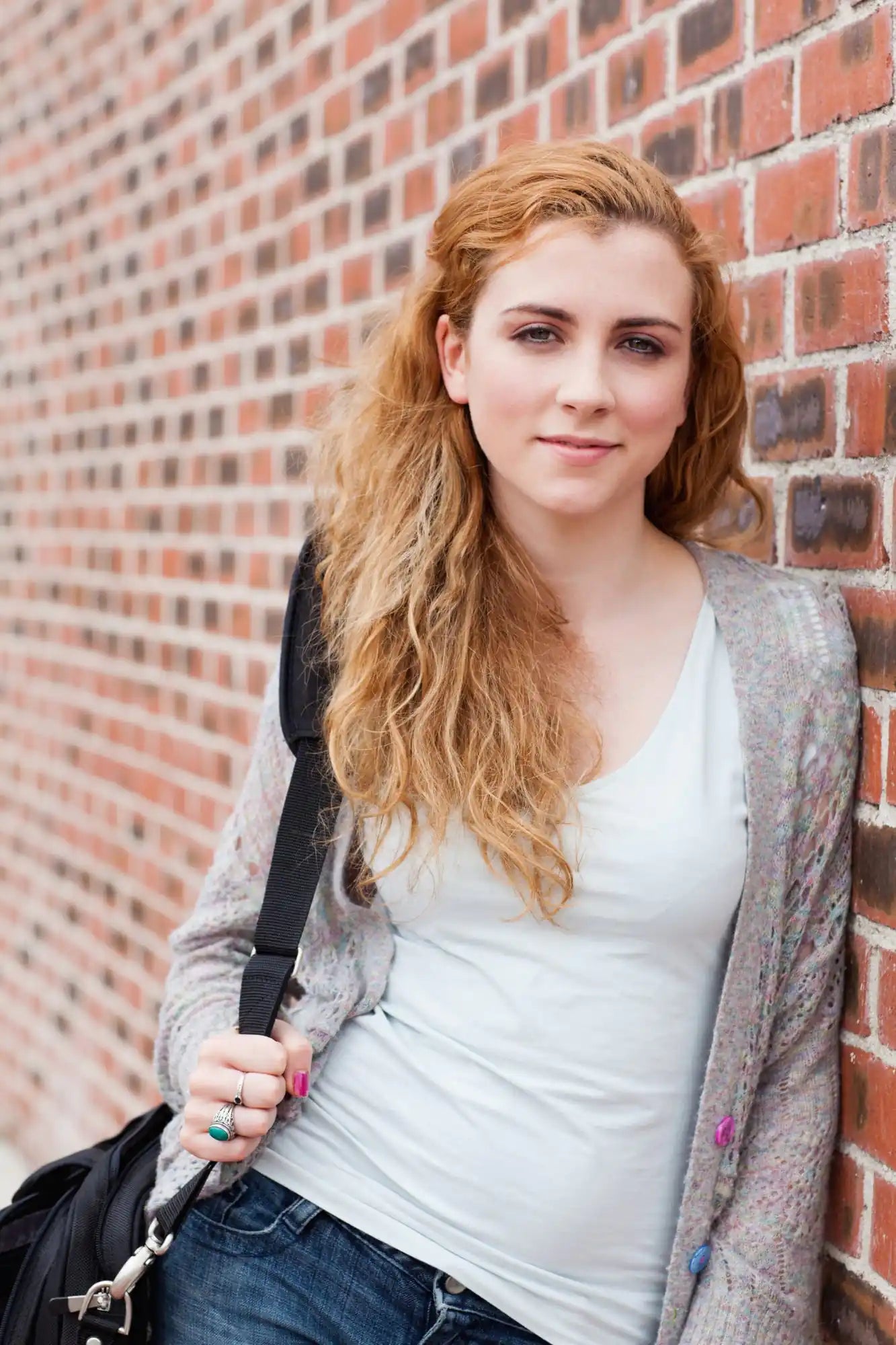 Woman with long auburn hair leans against a red brick wall, wearing a light top and cardigan.