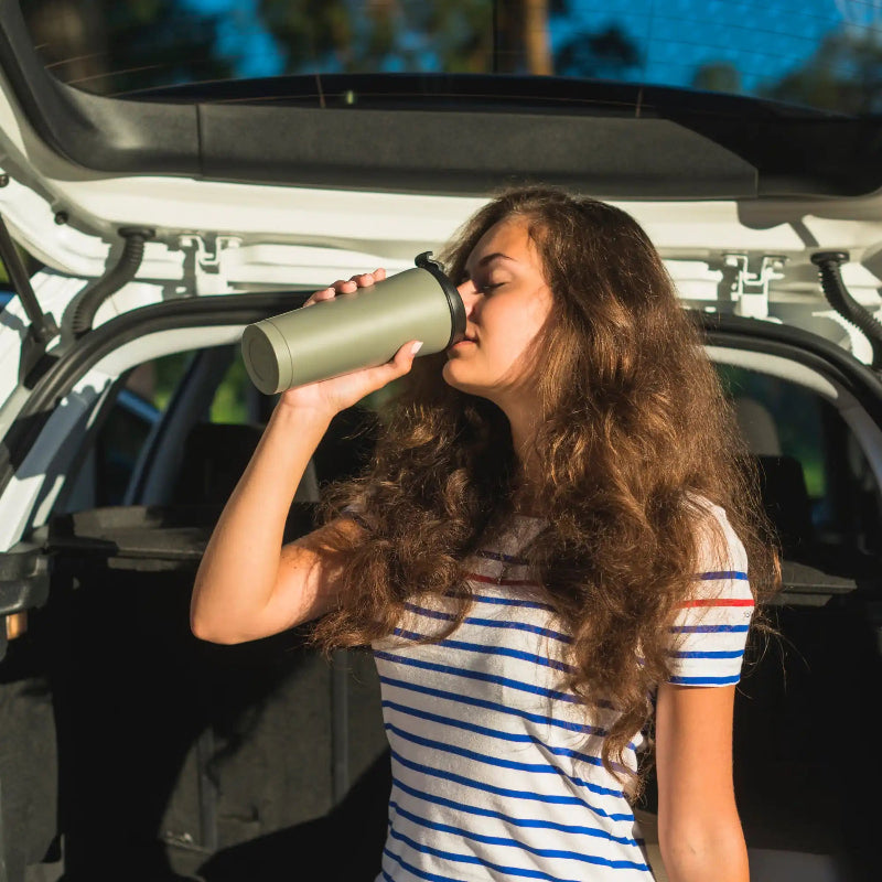 Woman in a car seat drinks from a gray insulated bottle, eyes closed, striped shirt visible.