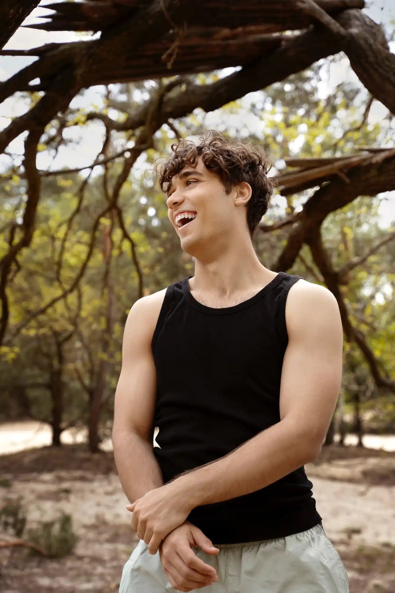 Young man with curly hair smiles in a black tank top outdoors under tree branches with golden leaves.