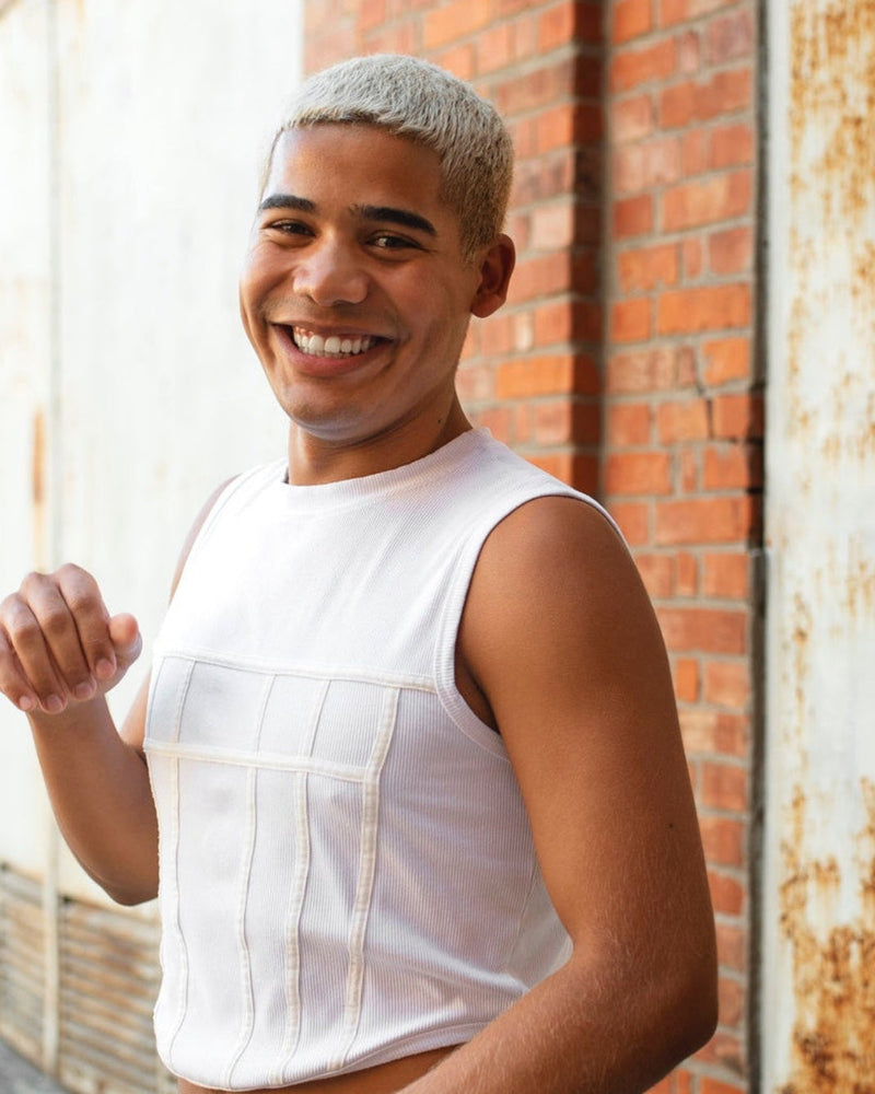 Smiling man in a white sleeveless shirt stands beside a red brick wall with rusty stains in warm light