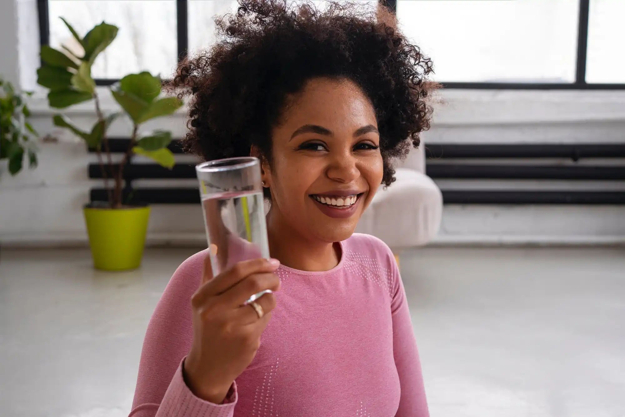 Smiling woman with curly hair holds a clear drinking glass, wearing a pink top in a bright room.