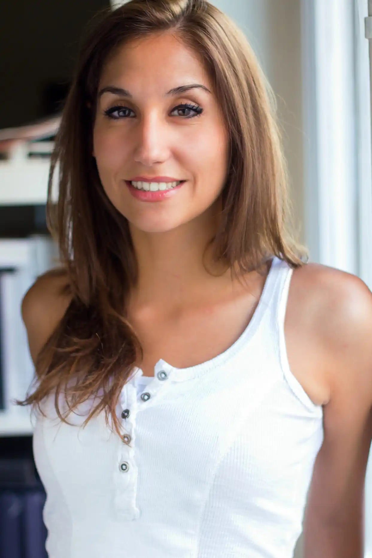 Smiling woman with long brown hair in a white sleeveless top, lit by soft window light indoors.