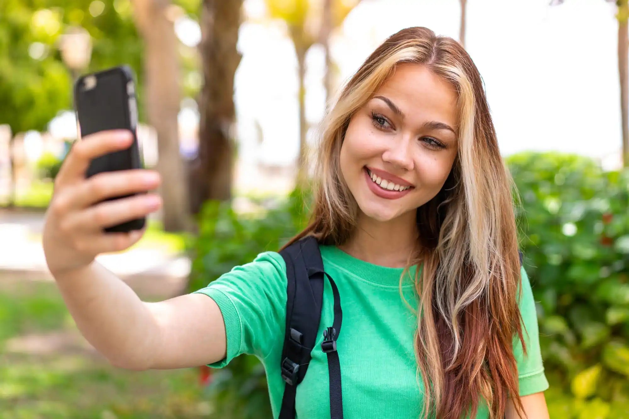 Woman taking a selfie with a black smartphone in a green garden, wearing a bright green shirt and backpack.