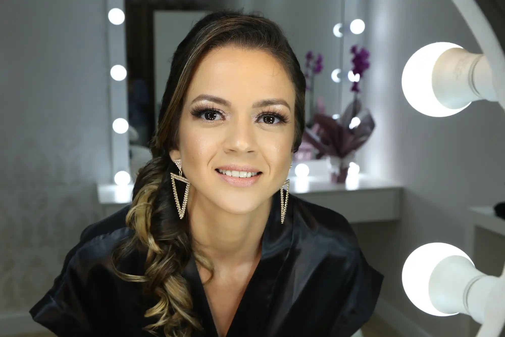 Smiling woman in a beauty studio with gold earrings, framed by mirror ring lights and blurred decor.