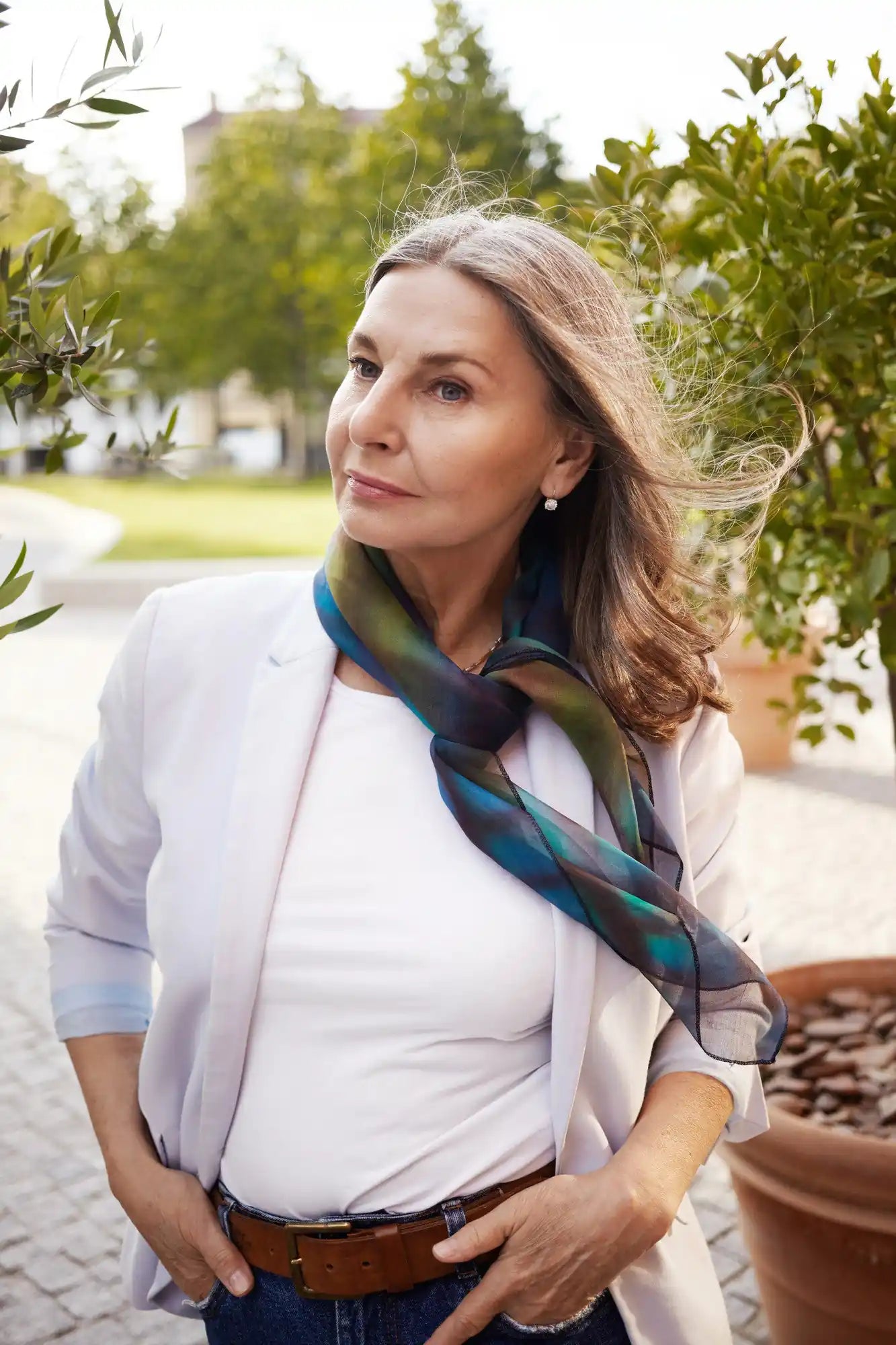 Woman in white blazer outdoors, wearing a blue-green patterned scarf as wind lifts her hair in a garden.