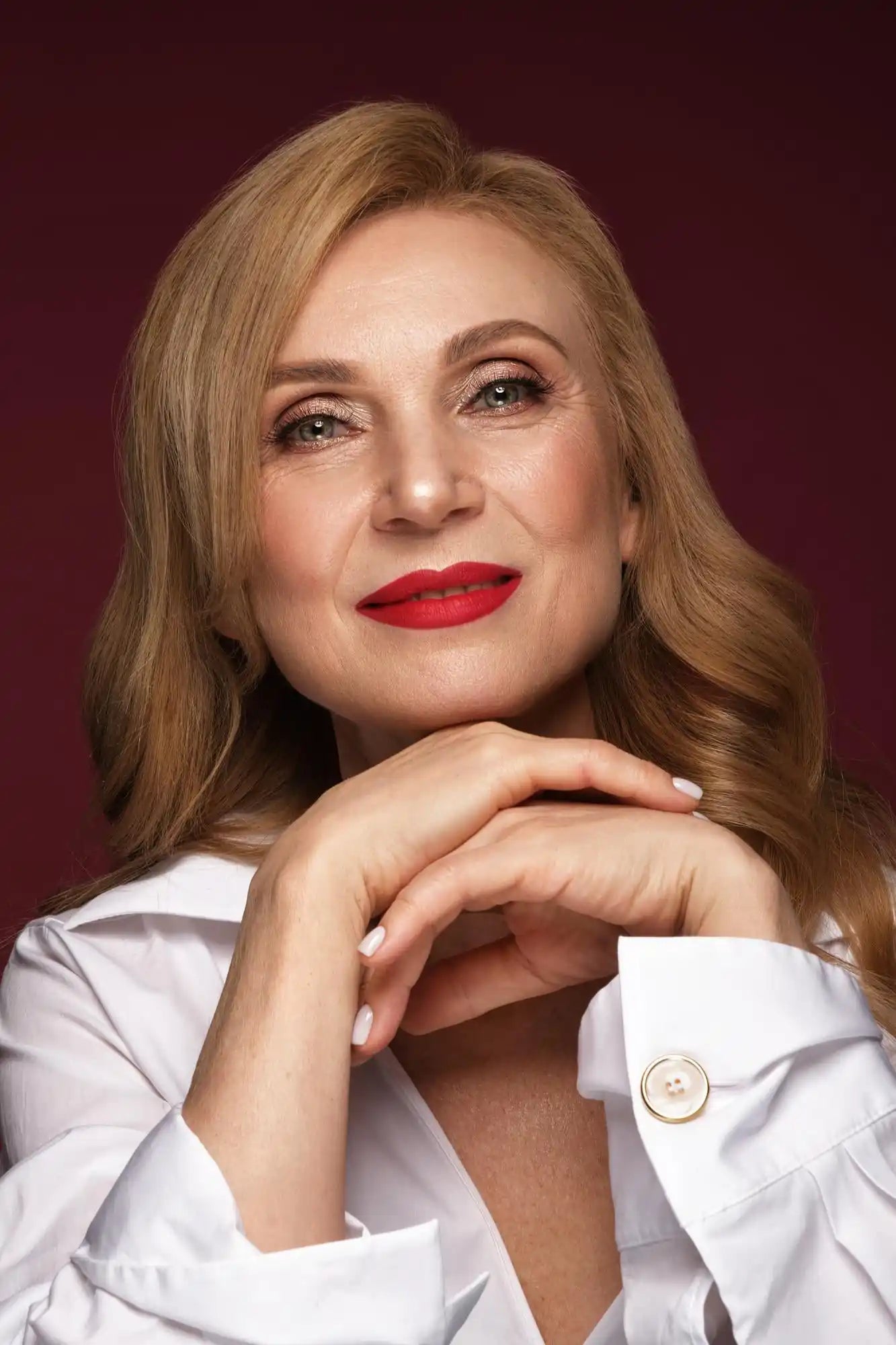 Woman portrait with long blonde hair, red lipstick, and hands clasped on a dark red studio backdrop.