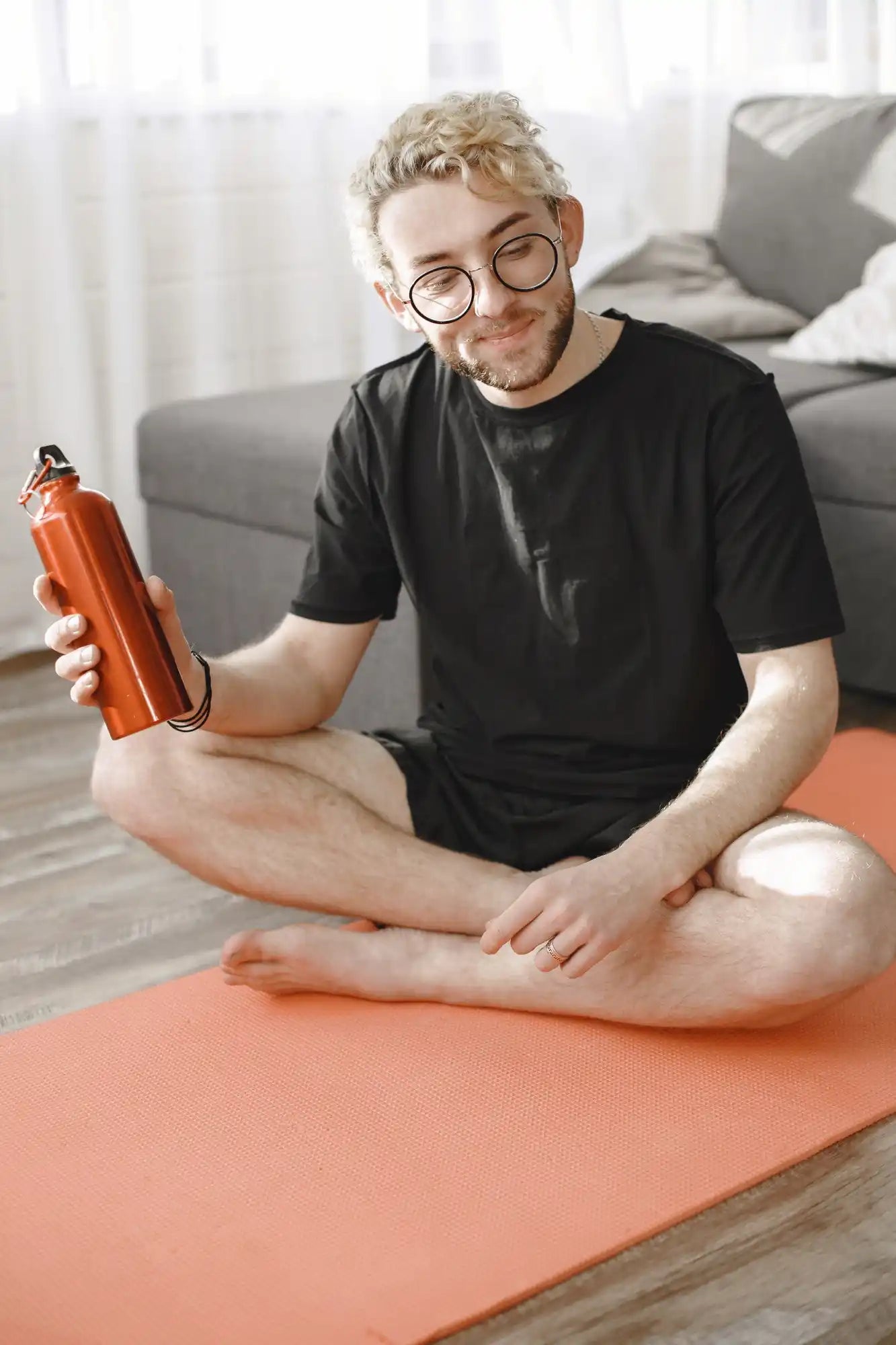 A man with glasses sits on an orange exercise mat on a couch, holding an orange water bottle.