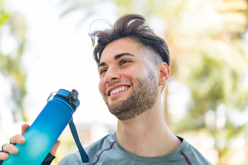 A smiling man holds a blue camera on a strap, selfie-style, with a sunlit garden blurred behind him.