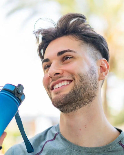 A smiling man holds a blue camera on a strap, selfie-style, with a sunlit garden blurred behind him.