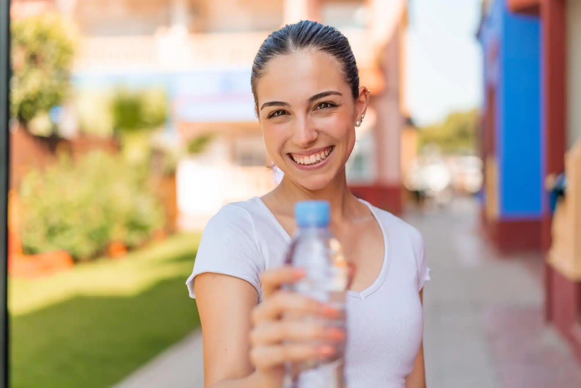 Smiling woman in a pale shirt holds a clear bottle in a sunny outdoor courtyard with blurred buildings.