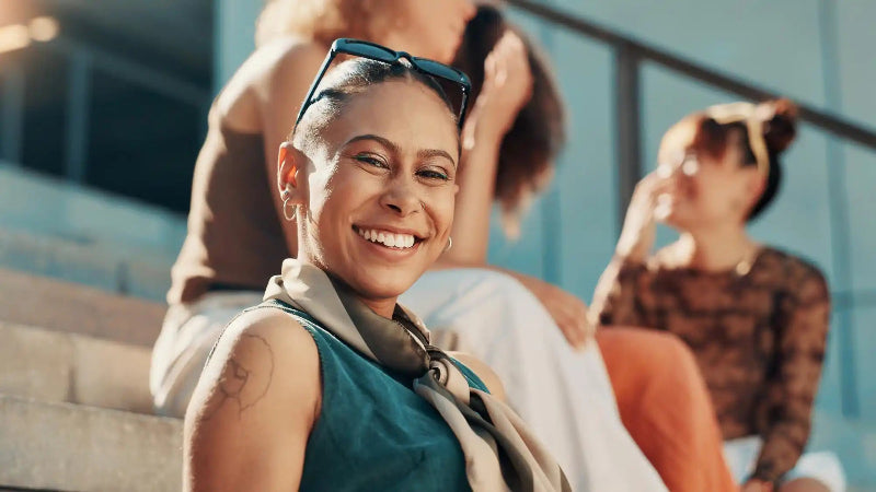 Smiling woman in teal vest with sunglasses on head relaxes outdoors as others blur behind her.