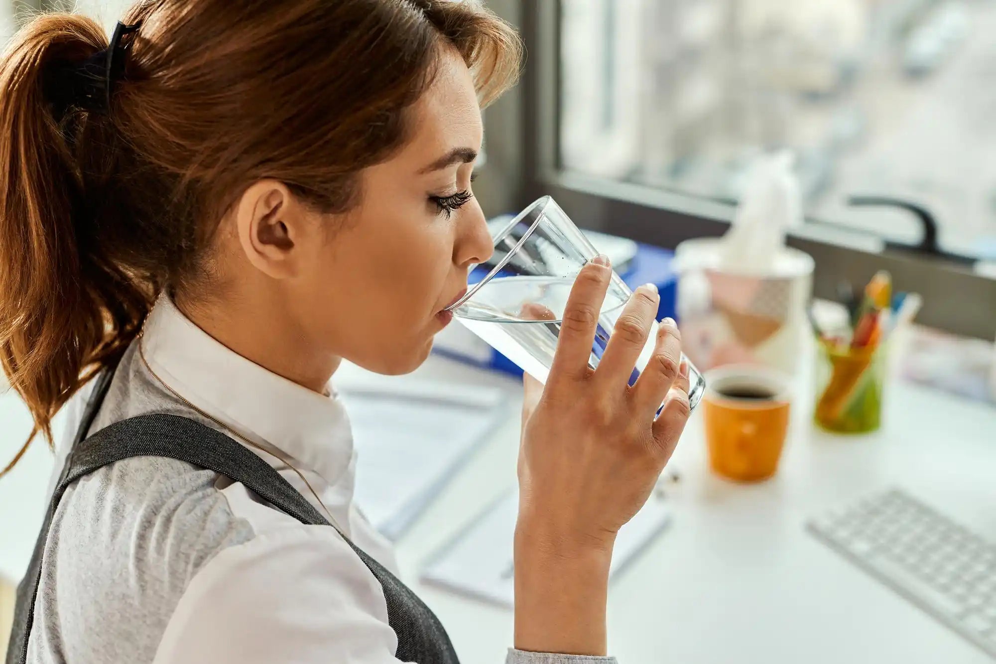 Woman in profile sips water from a clear glass, with a blurred home kitchen background.