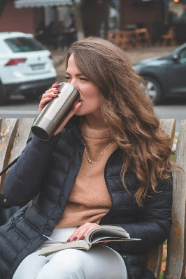 Woman on a wooden bench drinks from a stainless steel bottle, with parked cars blurred behind her.