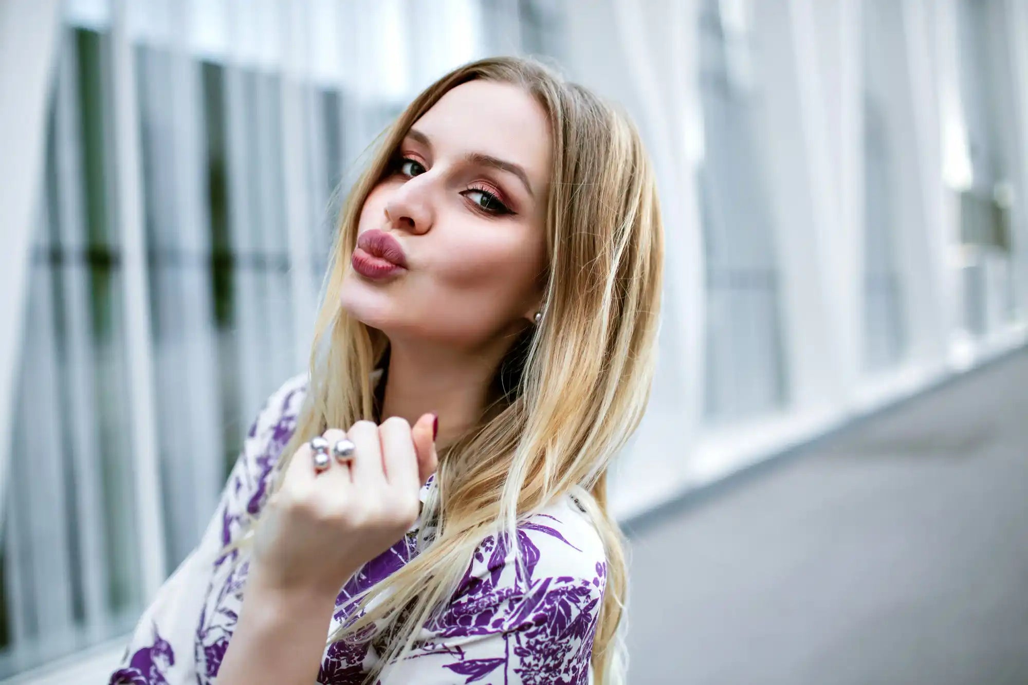 Blonde woman in a purple patterned top poses outdoors, lips pursed and rings visible against a blurred building.