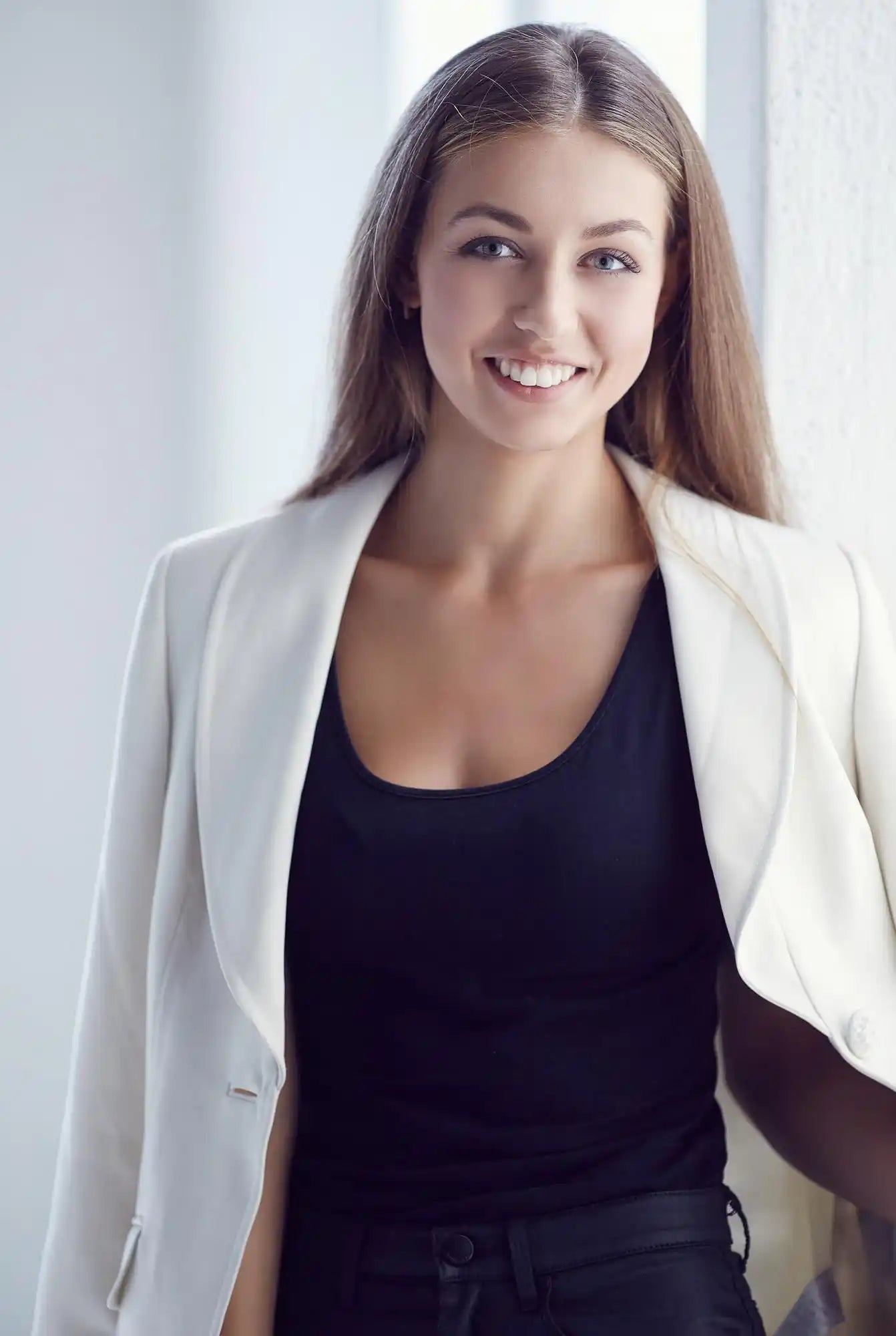 A smiling woman with straight brown hair wears a white blazer over a dark top against a light background.