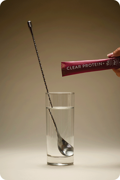 Hand squeezes a red CLEAR PROTEIN+ packet into a clear water glass while a metal rod stirs.