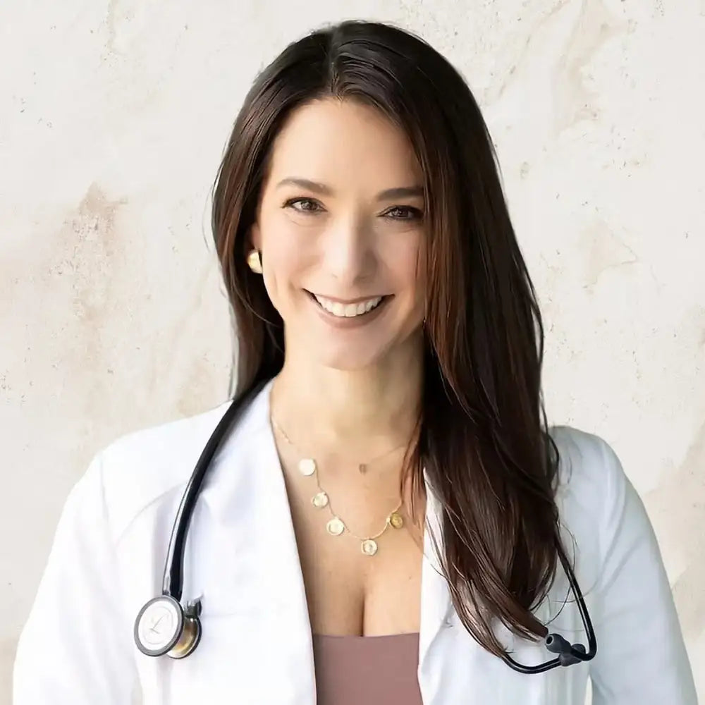 Smiling doctor in a white lab coat with stethoscope, dark hair, and gold necklace against a pale wall.