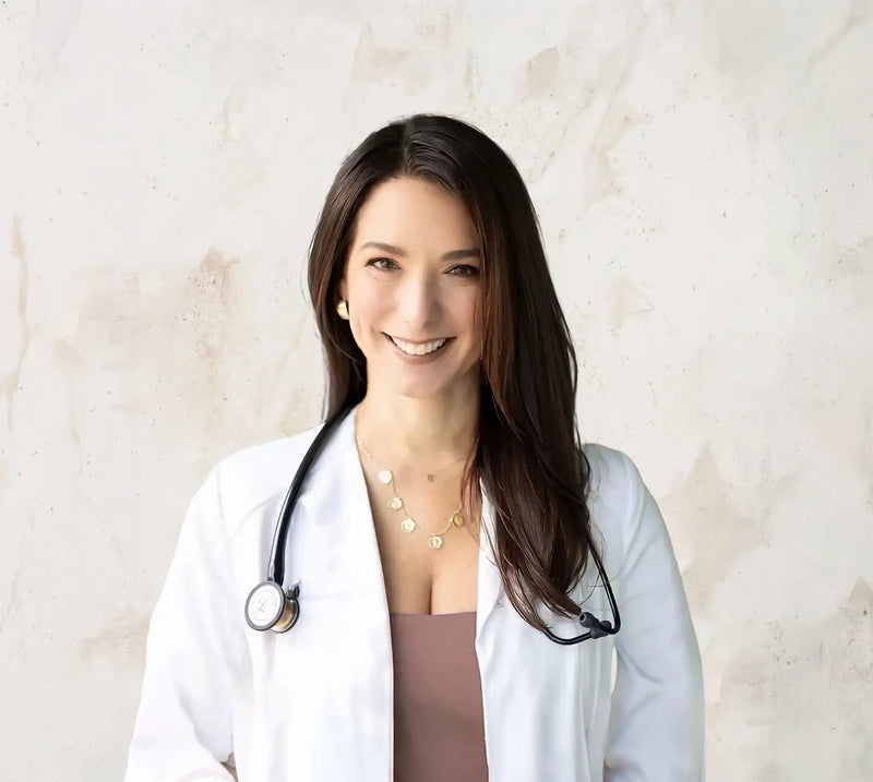 Smiling female doctor with long dark hair wears a white coat and stethoscope against a light wall.