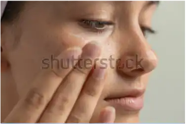 Woman applying skincare cream to her cheek and under eye, with fingers and soft lighting on skin