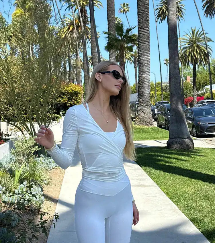 Woman in white top and trousers, wearing sunglasses, stands on a sunny garden walkway beside palm trees.