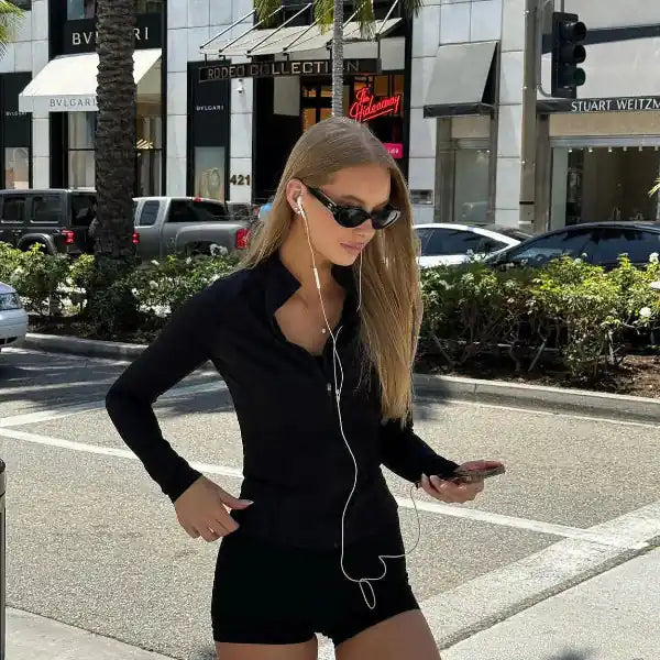 Woman in black outfit and sunglasses walks on a city street, long blonde hair with white earphones.