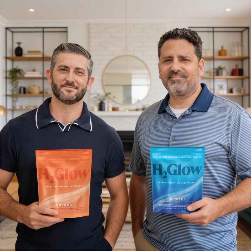 Two men in a bright kitchen holding H2Glow electrolyte pouches, one orange and one blue