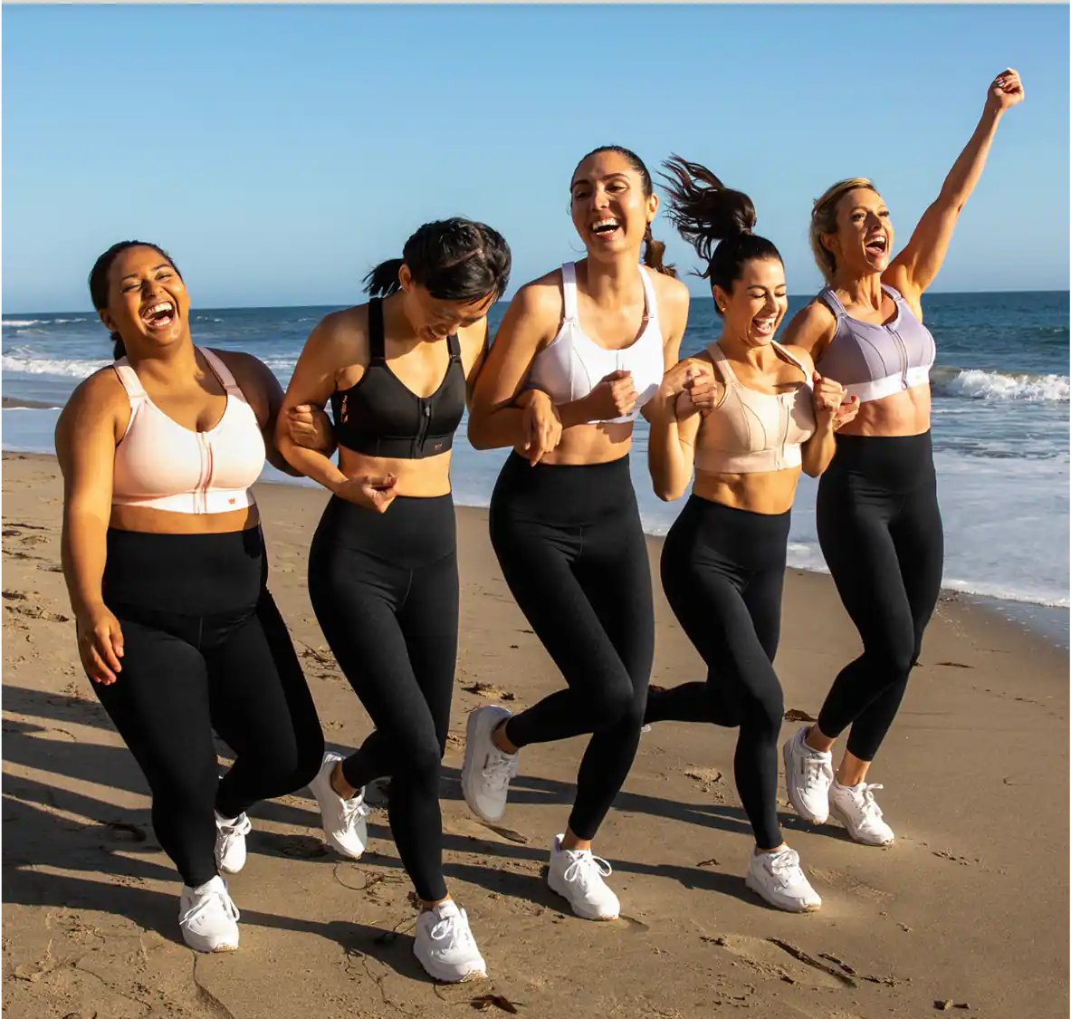 Five women jog along a sandy beach in white and black leggings, laughing with arms raised against the ocean.