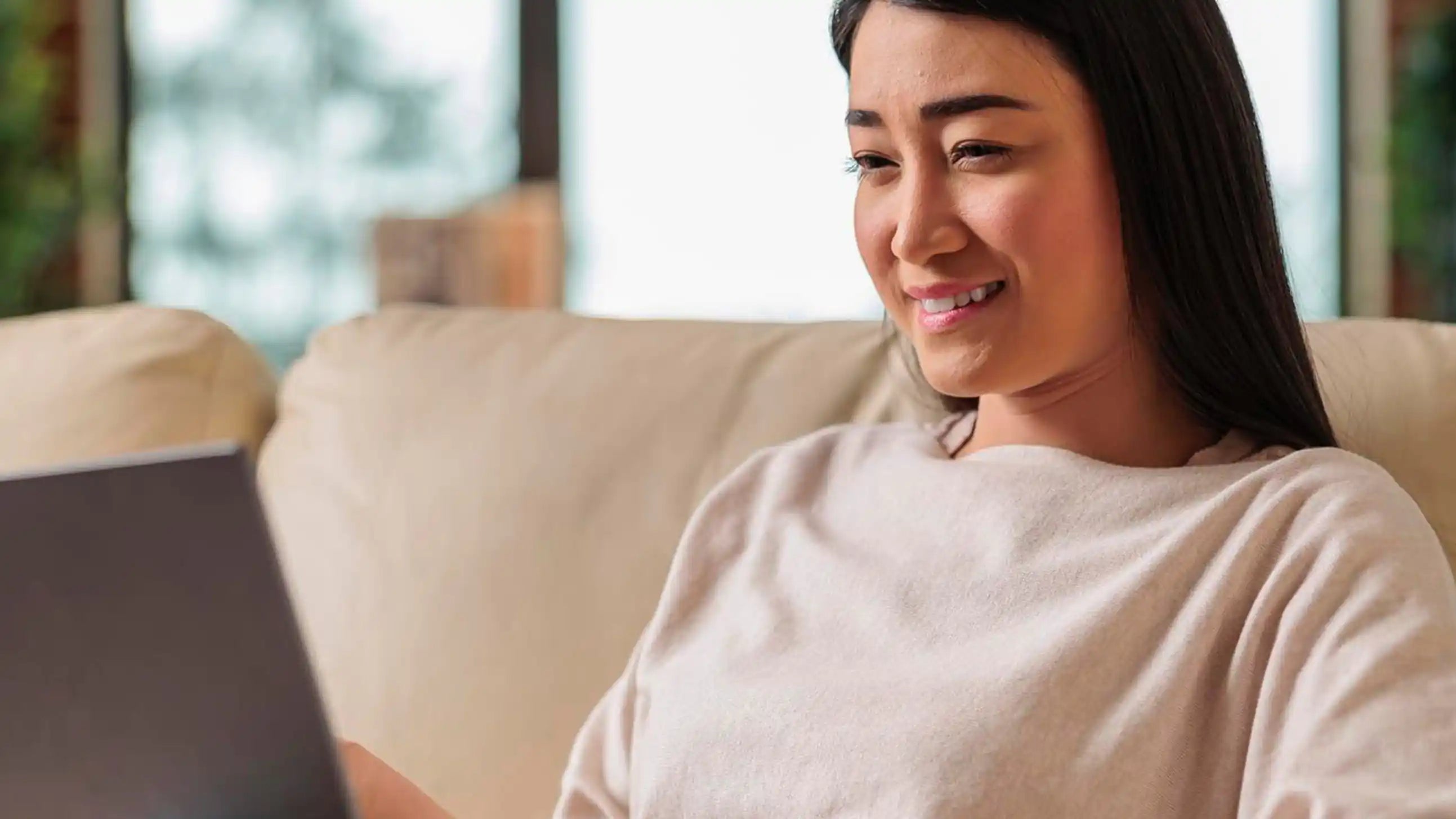 Woman reclining on a beige sofa, wrapped in a light blanket, smiling at an open laptop screen.