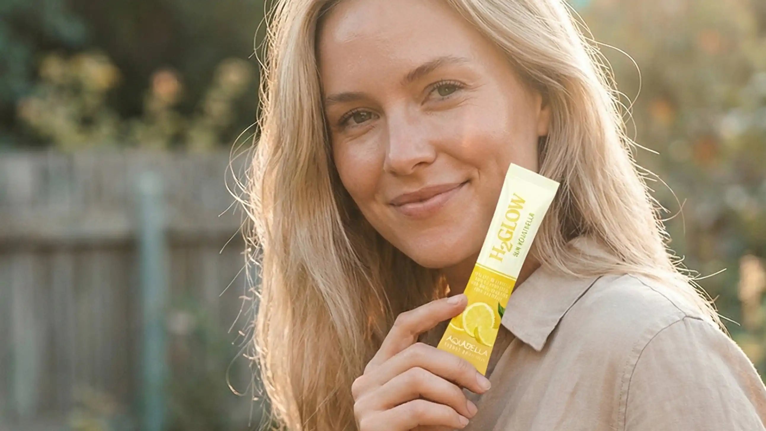 Smiling woman with long blonde hair holds a yellow and white stick packet outdoors in soft sunlight