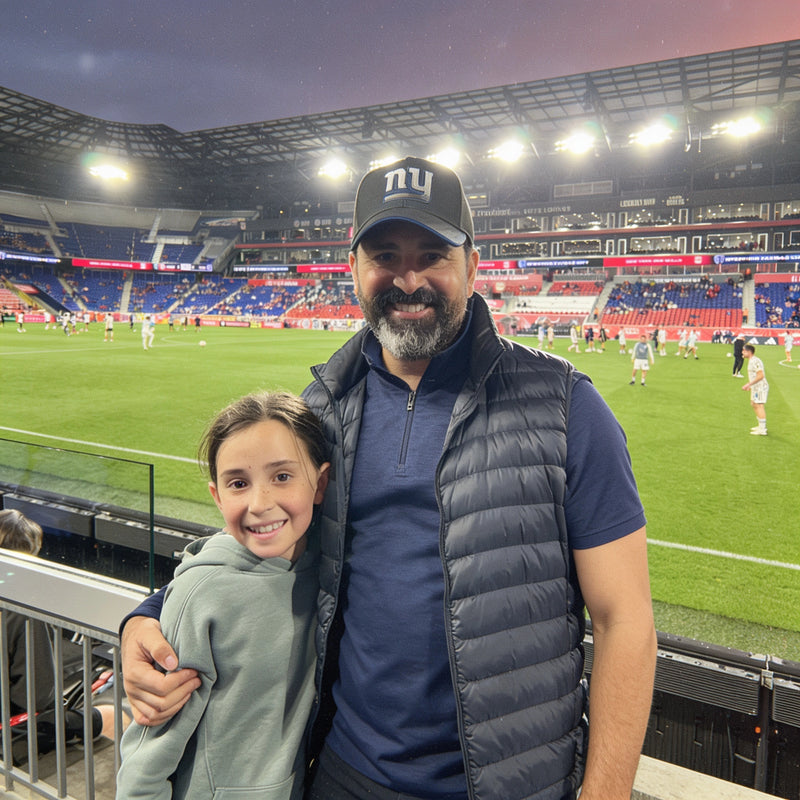 Man in a dark cap and puffy vest at a soccer stadium, with a child in front and bright lights behind