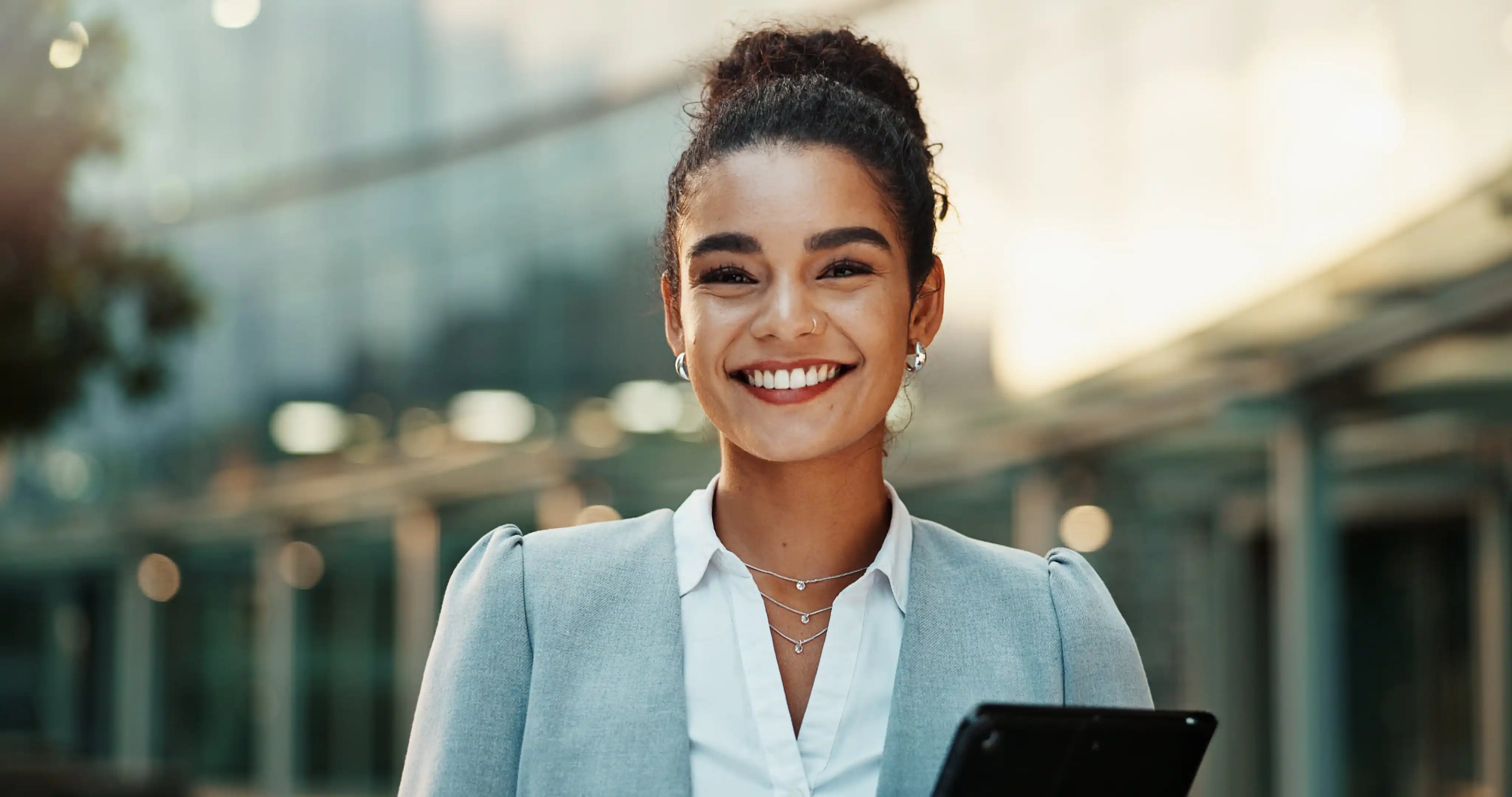 Smiling woman in a light blue blazer holds a tablet, with a softly lit cafe background behind her.