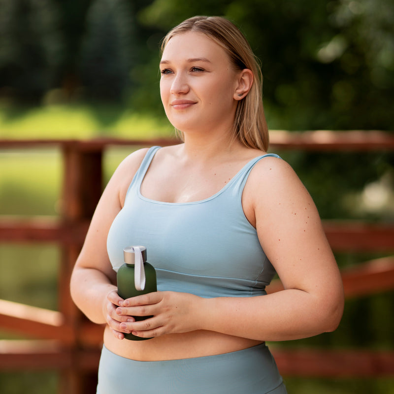 Woman in a light blue tank top holding a green bottle outdoors, smiling with blond hair tied back