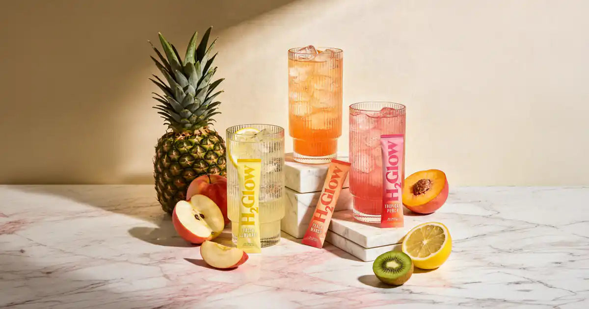 Pineapple and sliced fruits sit beside tall red, pink, yellow, and clear beverage cans on a marble counter.