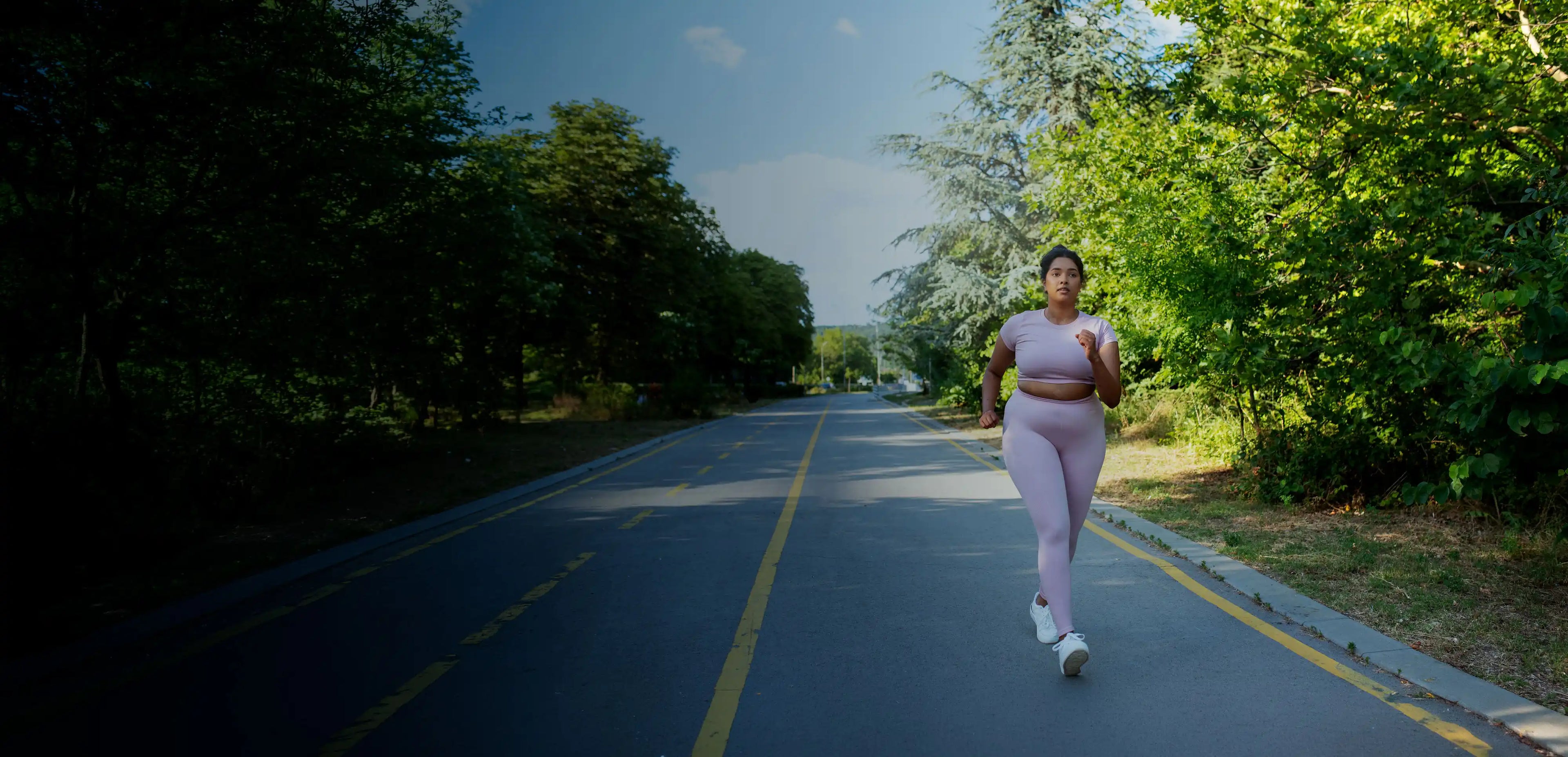 A woman jogs along a paved park road in light pink activewear, with green trees under bright sky.
