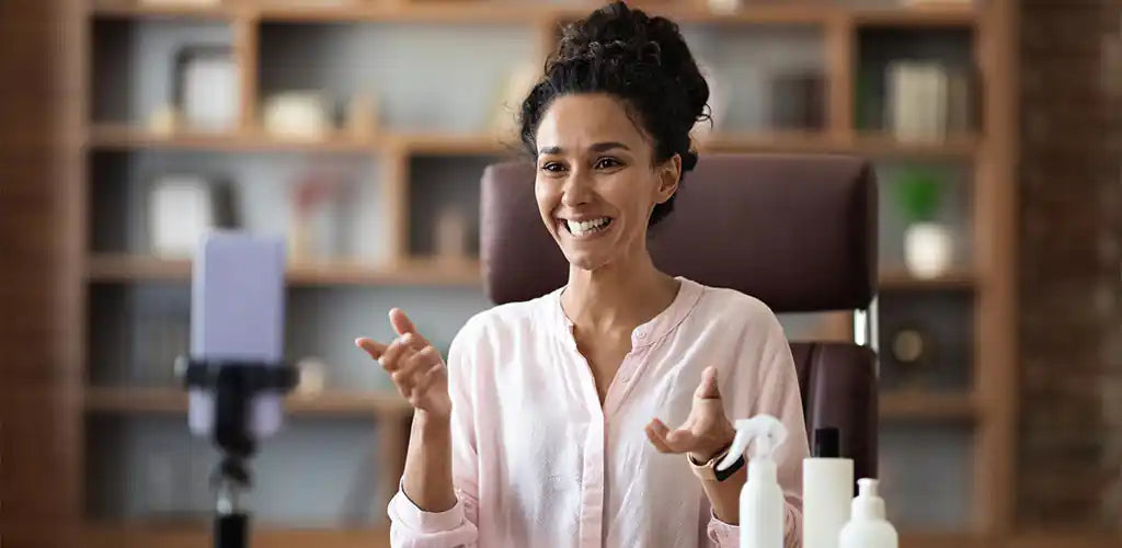 Smiling woman in a pale pink blouse gestures while seated at a desk with bottles in view.