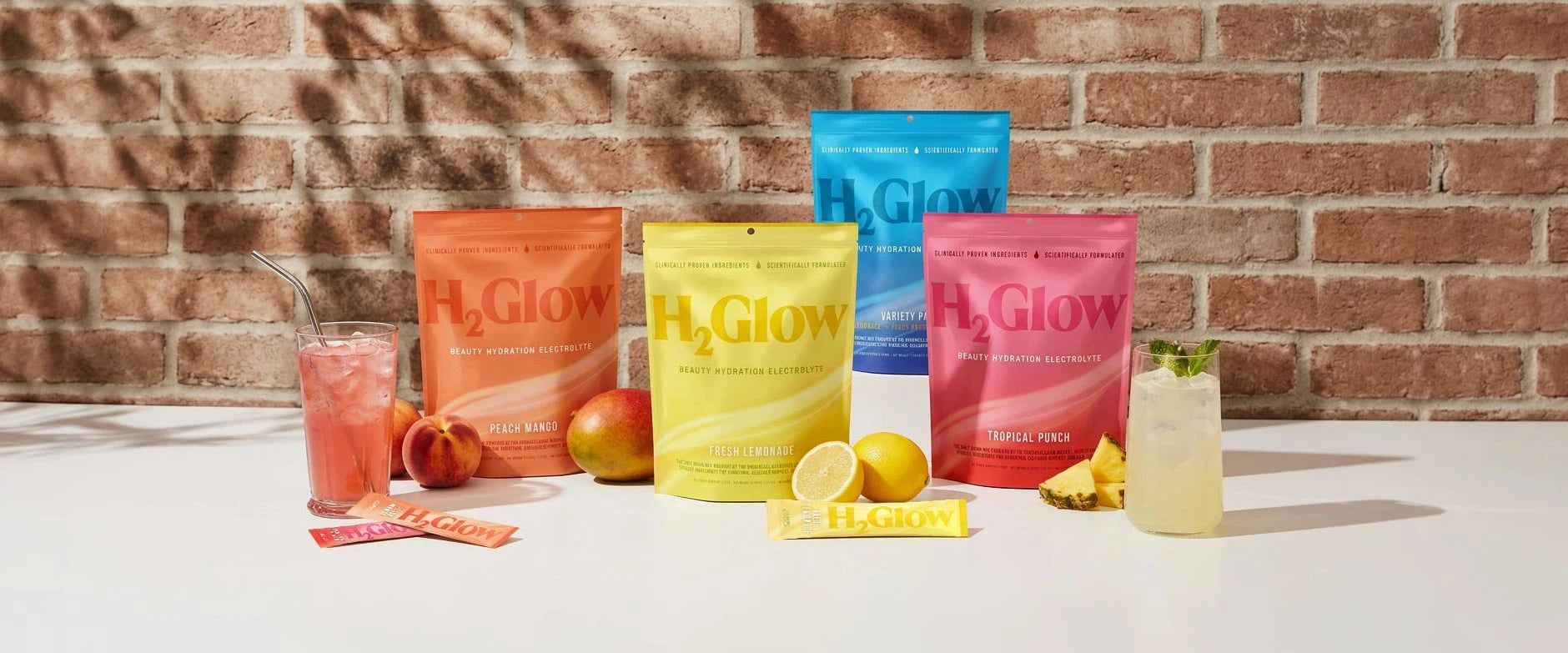 Colorful H2Glow electrolyte packets, with peach mango, fresh lemonade, and tropical punch sachets, beside drinks.